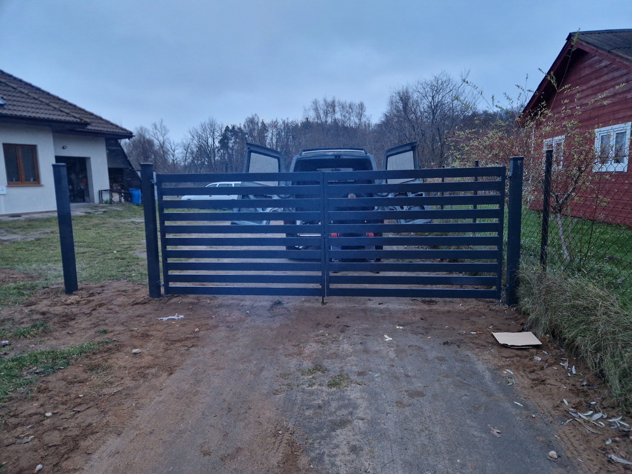 Nowoczesna, pozioma brama wjazdowa z metalowymi sztachetami w kolorze grafitowym, zamontowana na posesji z widocznym samochodem dostawczym w tle.