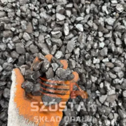 Dłoń w roboczej rękawicy trzyma bryłki węgla na tle hałdy węgla. Widoczne logo firmy 'Szóstak Skład Opału'.