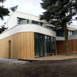 Oleksandr Otinov Aleks Tarasy - Nowoczesny dom z elewacją z jasnego drewna, dużymi oknami i balkonami z szklanymi balustradami, otoczony drzewami, w trakcie prac wykończeniowych tarasu.