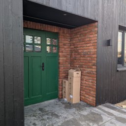 Oleksandr Otinov Aleks Tarasy - Nowoczesne wejście do budynku z ciemną, drewnianą elewacją i zielonymi drzwiami. Widoczna ceglana ściana i stos kartonów obok wejścia.