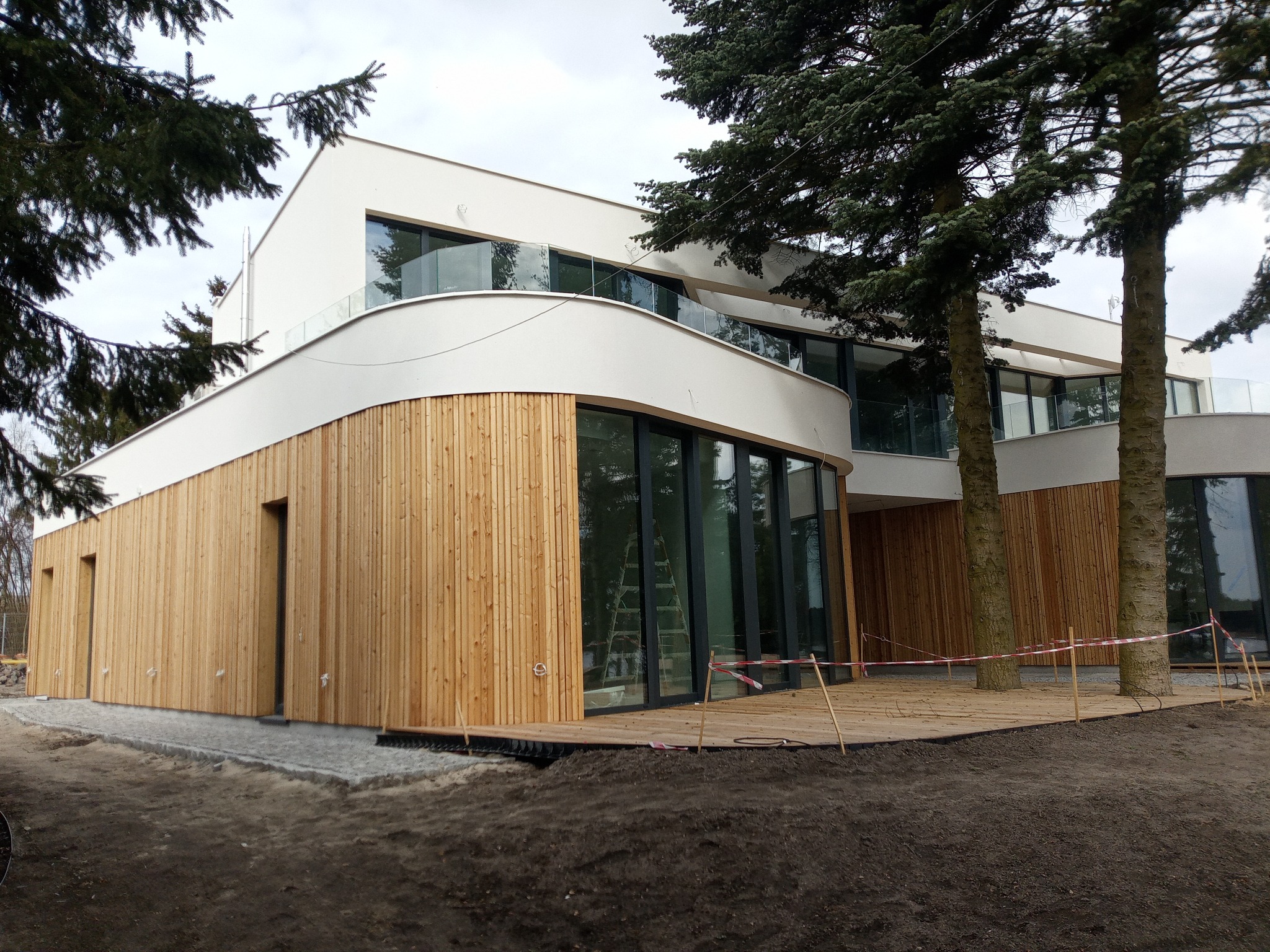 Nowoczesny dom z elewacją z jasnego drewna, dużymi oknami i balkonami z szklanymi balustradami, otoczony drzewami, w trakcie prac wykończeniowych tarasu.