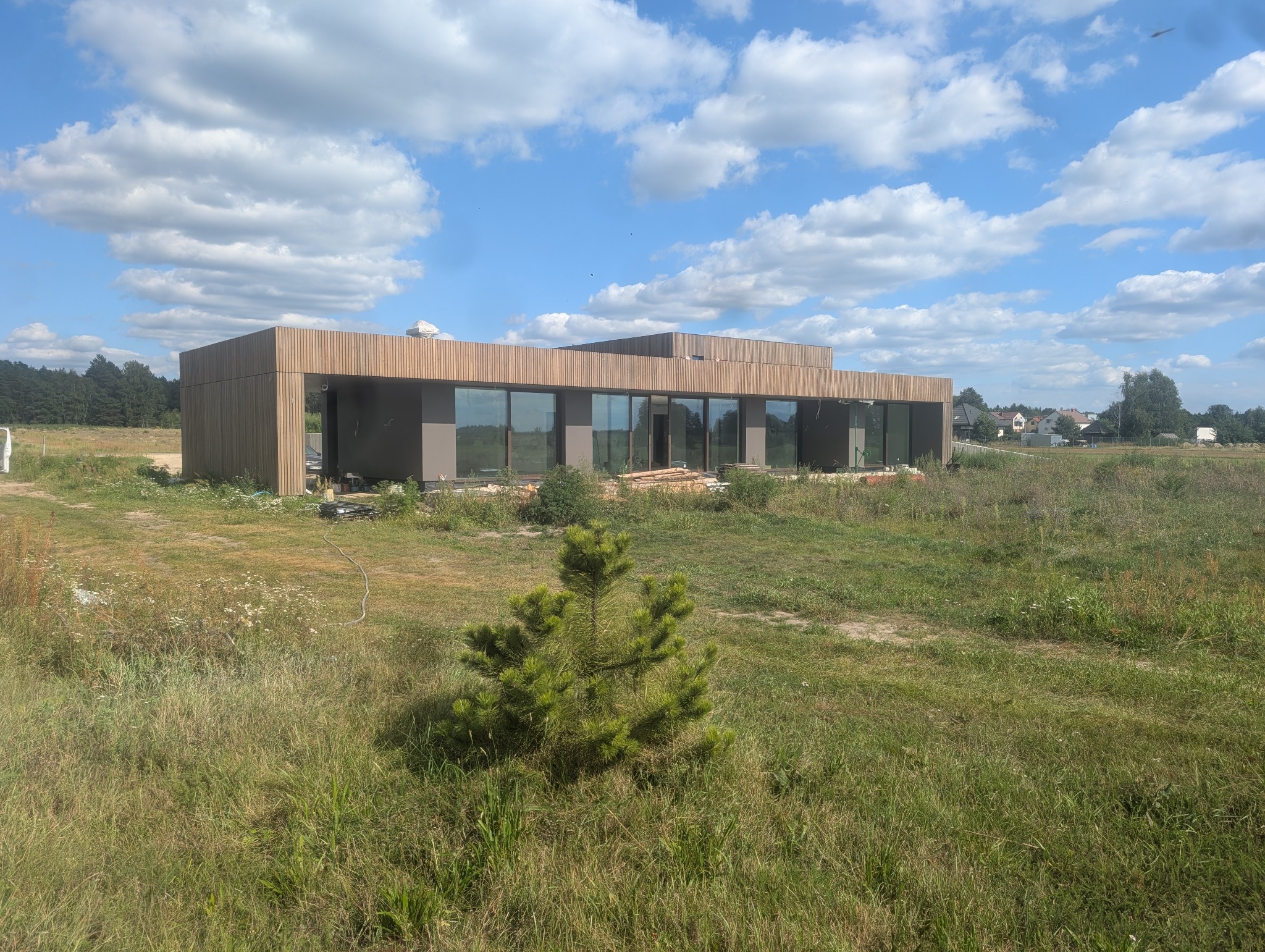 Nowoczesny dom jednorodzinny z drewnianą elewacją i dużymi oknami, otoczony zielenią. Widoczne elementy wykończeniowe i przygotowanie terenu.