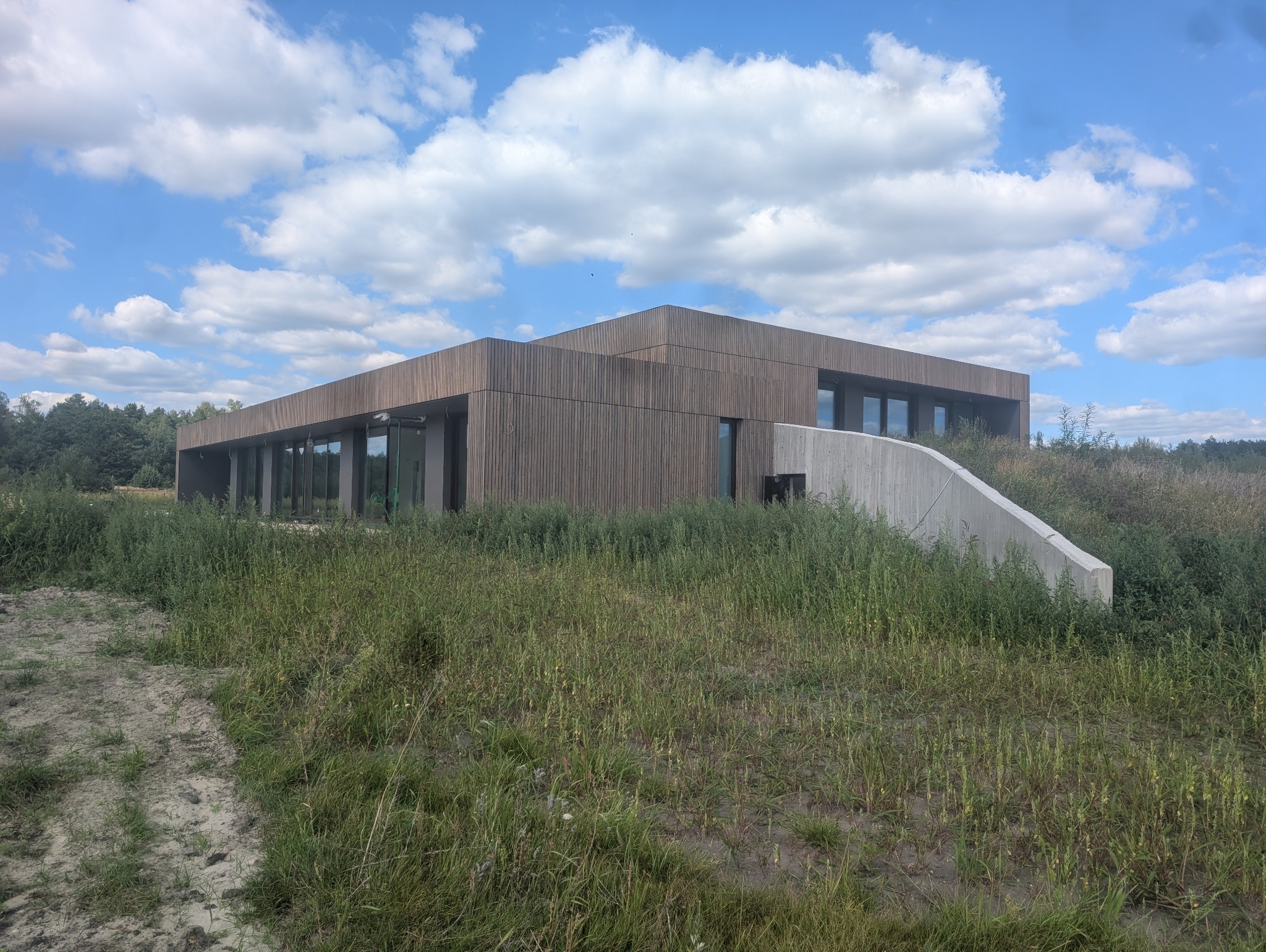 Nowoczesny budynek z drewnianą elewacją i betonowym elementem, otoczony zielenią. Widoczne duże okna i błękitne niebo z chmurami.