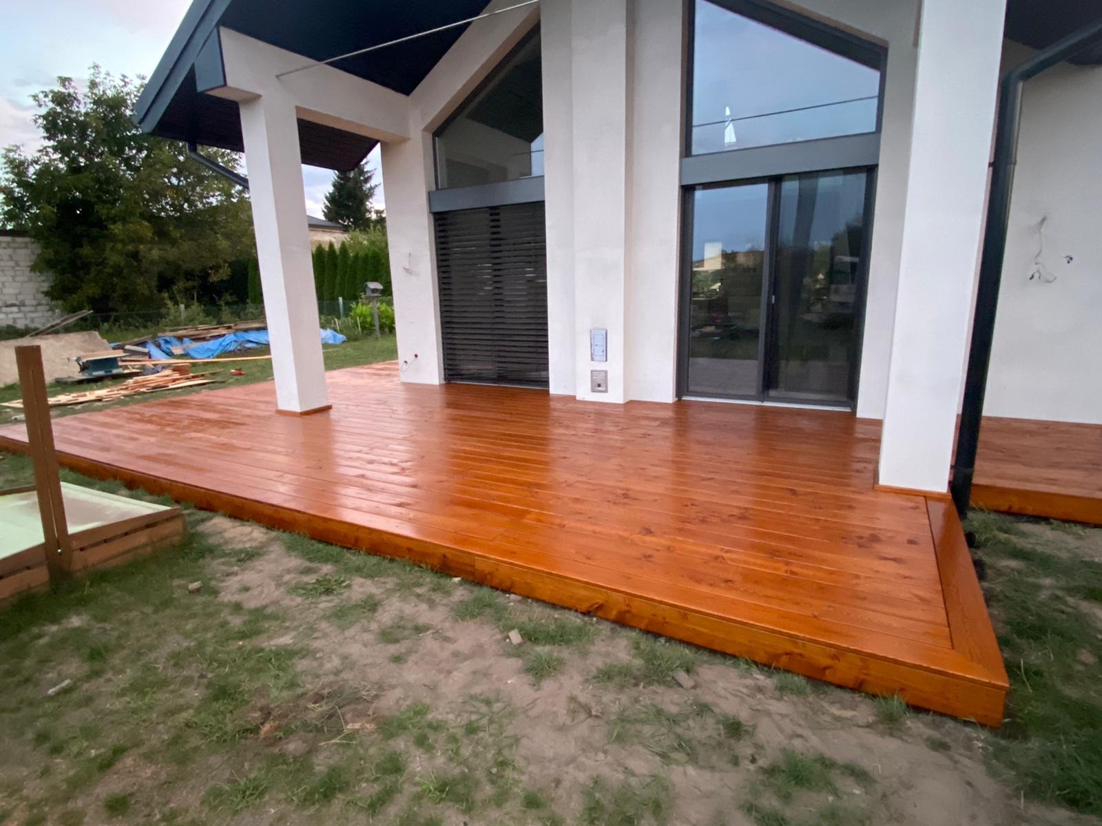 Nowoczesny taras drewniany z desek w kolorze ciepłego brązu, przylegający do białego budynku z dużymi oknami. Widoczne elementy konstrukcyjne i trawa.