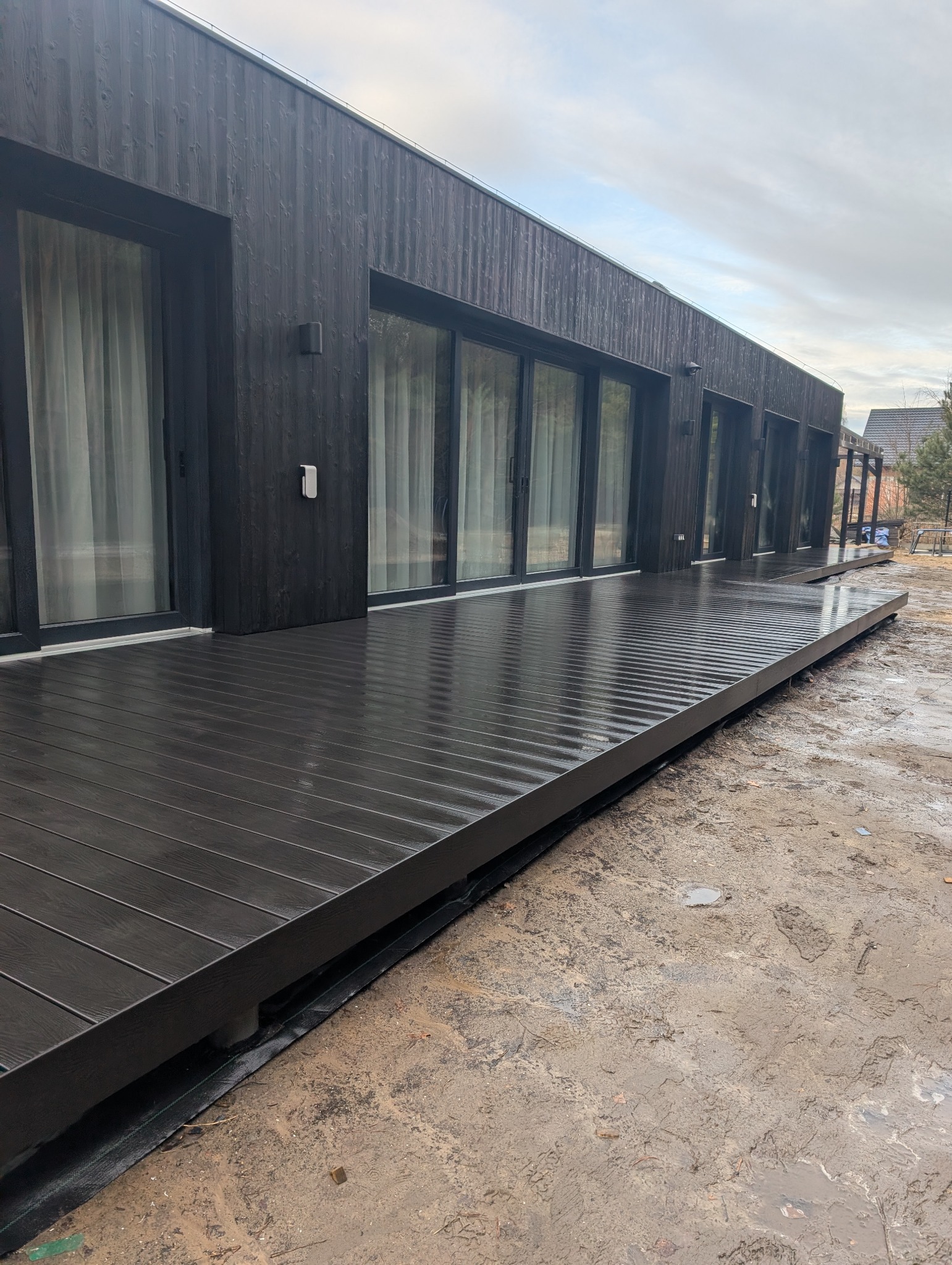 Nowoczesny taras kompozytowy przy domu z ciemną elewacją i dużymi oknami przesuwnymi, widok z boku na mokrą powierzchnię tarasu po deszczu.