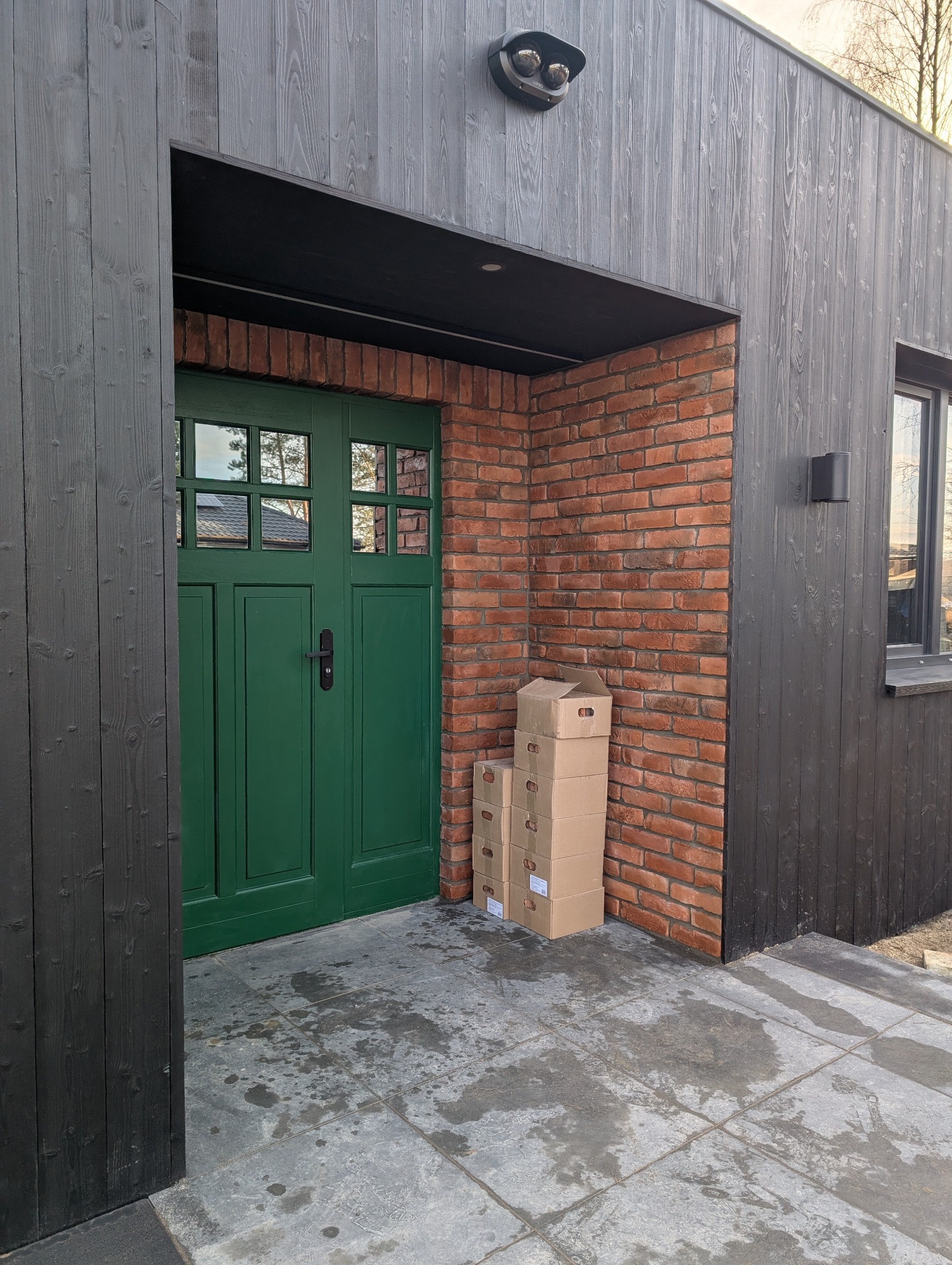 Nowoczesne wejście do budynku z ciemną, drewnianą elewacją i zielonymi drzwiami. Widoczna ceglana ściana i stos kartonów obok wejścia.
