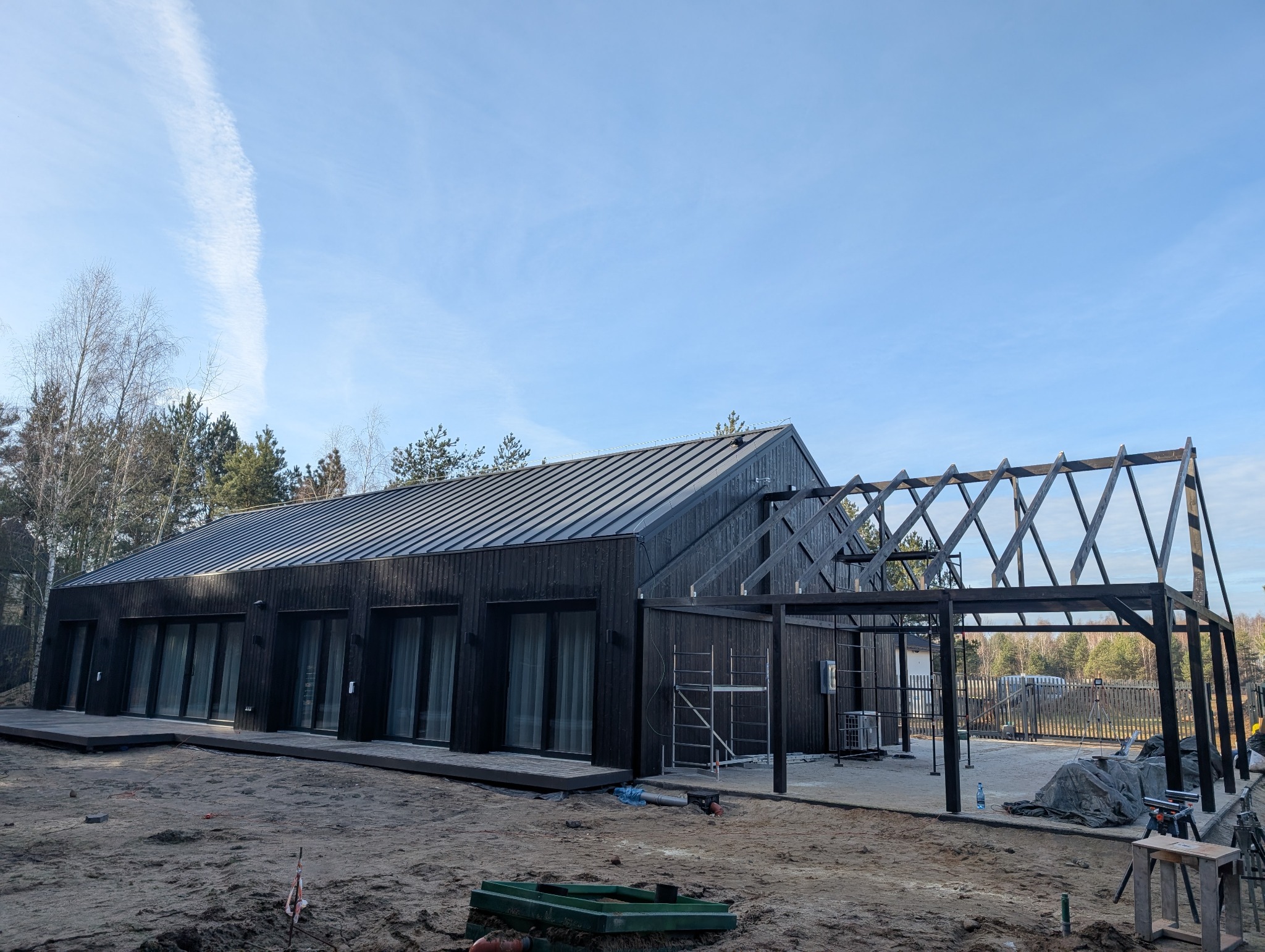 Nowoczesny dom z ciemnego drewna z metalowym dachem i drewnianą konstrukcją tarasu w trakcie budowy, na tle lasu i błękitnego nieba.