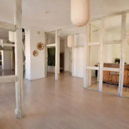 Jasne, przestronne wnętrze sali eventowej z drewnianą podłogą, dekoracyjnymi lampami i barem z rzeźbionym frontem. Widoczne drewniane konstrukcje i elementy dekoracyjne.