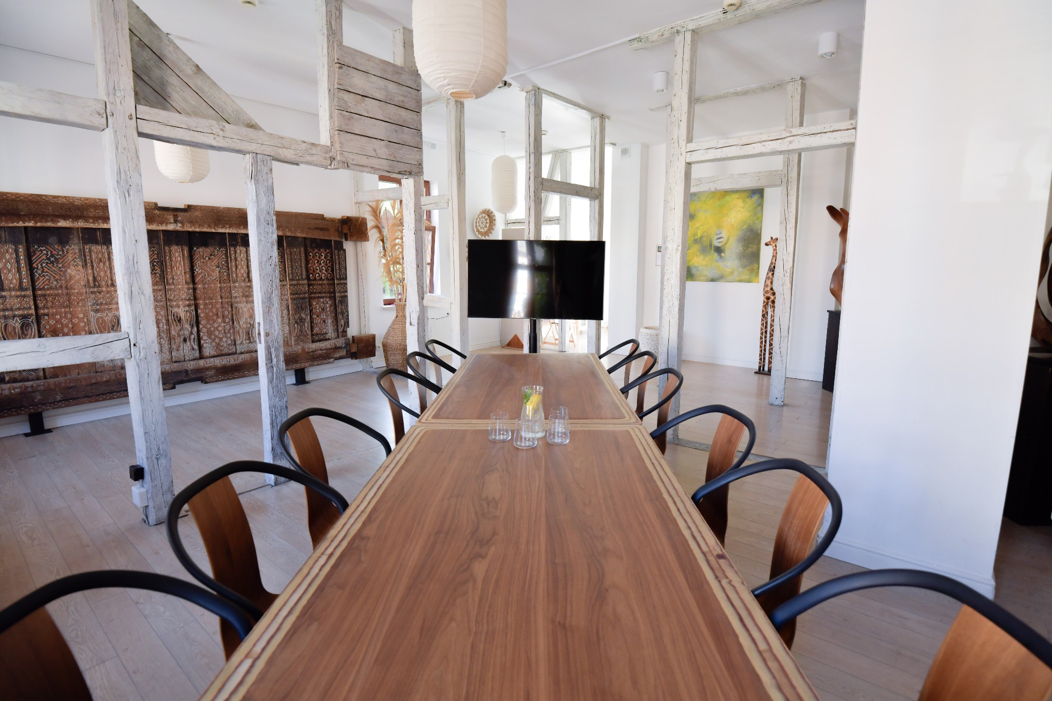 Sala konferencyjna z długim drewnianym stołem, krzesłami, i telewizorem. W tle dekoracyjne elementy architektoniczne i oświetlenie.