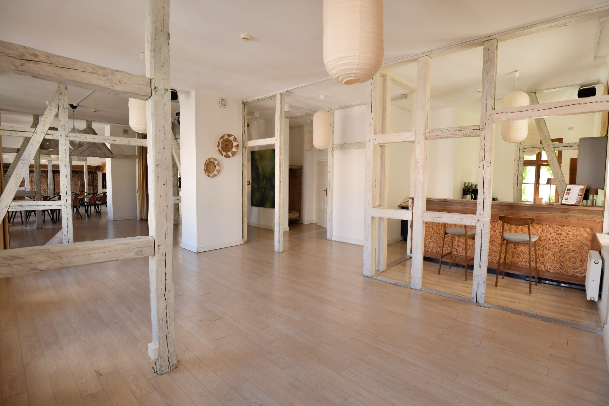 Jasne, przestronne wnętrze sali eventowej z drewnianą podłogą, dekoracyjnymi lampami i barem z rzeźbionym frontem. Widoczne drewniane konstrukcje i elementy dekoracyjne.