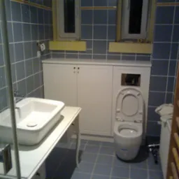 Łazienka z zabudową toalety i szafkami pod umywalką, niebieskie płytki na ścianach i podłodze, widoczny grzejnik drabinkowy.