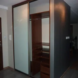 Szafa wnękowa z przesuwnymi drzwiami z mlecznego szkła, drewniane półki i szuflady, ciemnoszara ściana boczna, szare płytki podłogowe.