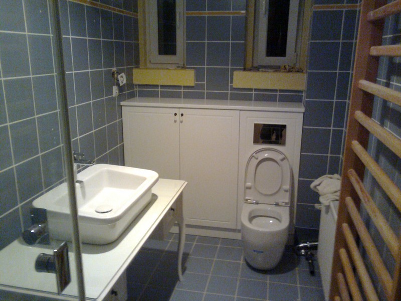 Łazienka z zabudową toalety i szafkami pod umywalką, niebieskie płytki na ścianach i podłodze, widoczny grzejnik drabinkowy.