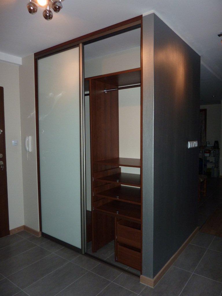 Szafa wnękowa z przesuwnymi drzwiami z mlecznego szkła, drewniane półki i szuflady, ciemnoszara ściana boczna, szare płytki podłogowe.