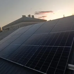 Panele fotowoltaiczne na dachu domu, widoczne mocowania i zachodzące słońce w tle. Instalacja w trakcie lub po montażu.