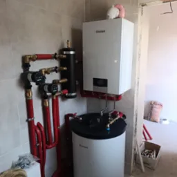 Nowoczesny, biały piec gazowy Vaillant z czerwonymi rurami, zainstalowany na tle szarych płytek. Widoczne elementy instalacji, w tym czarny zbiornik i pompy obiegowe.
