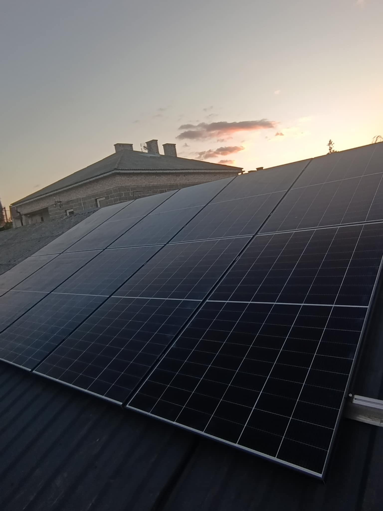 Panele fotowoltaiczne na dachu domu, widoczne mocowania i zachodzące słońce w tle. Instalacja w trakcie lub po montażu.