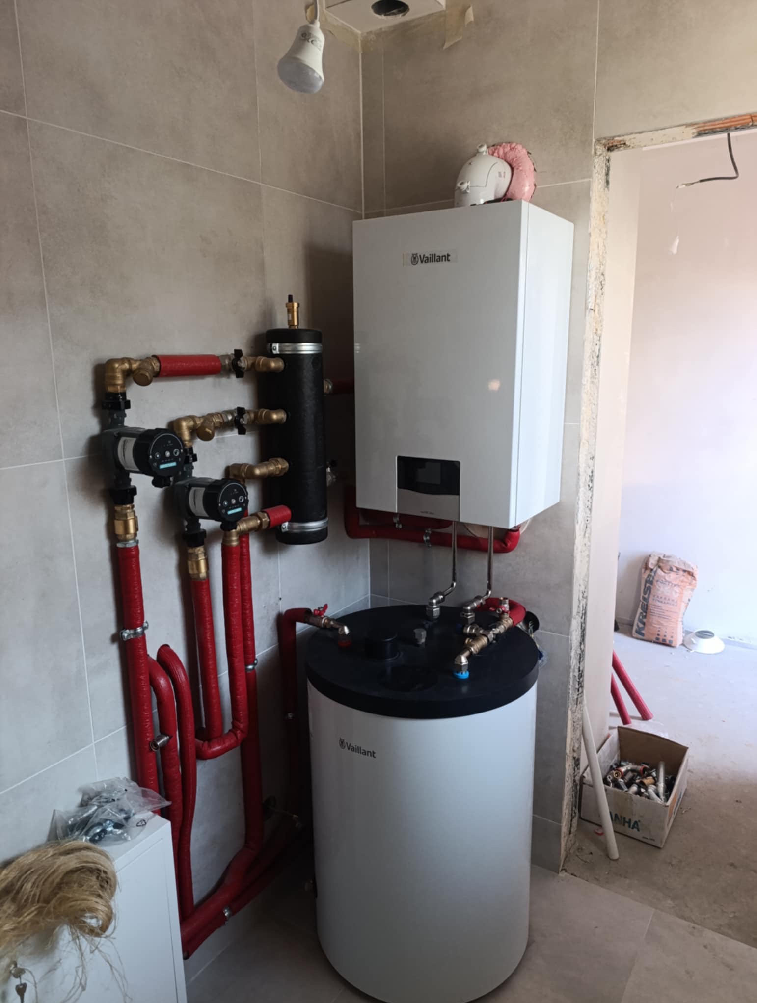 Nowoczesny, biały piec gazowy Vaillant z czerwonymi rurami, zainstalowany na tle szarych płytek. Widoczne elementy instalacji, w tym czarny zbiornik i pompy obiegowe.