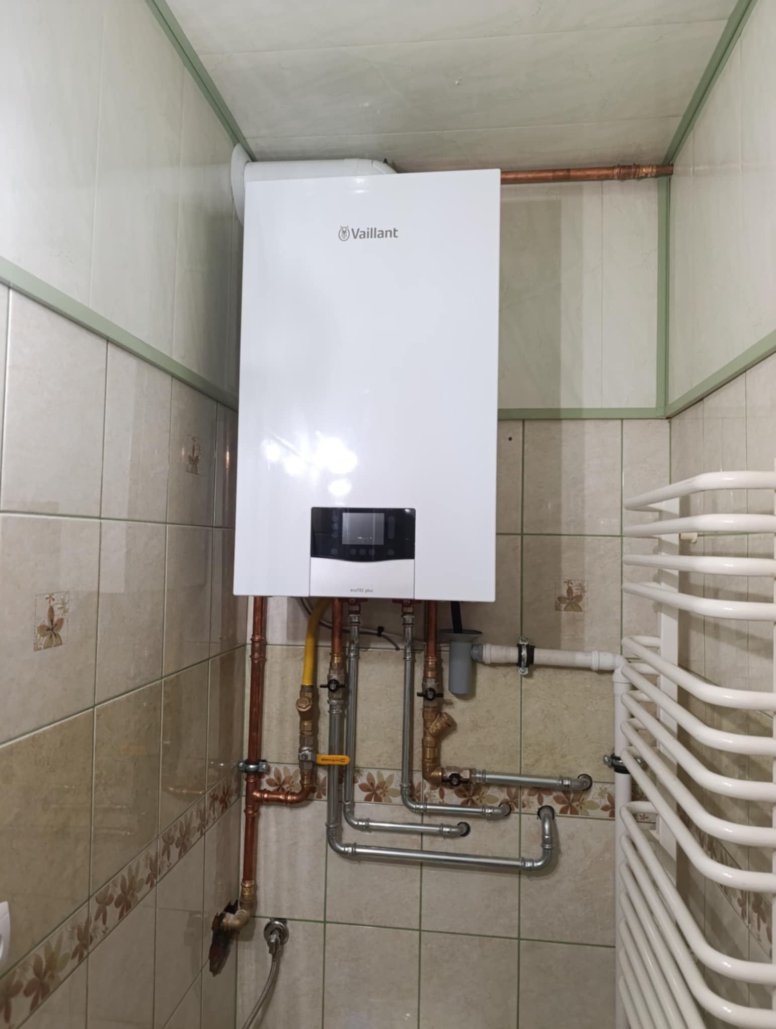 Biały, wiszący piec gazowy Vaillant w łazience z beżowymi płytkami, widoczne rury miedziane i grzejnik drabinkowy, instalacja po montażu.