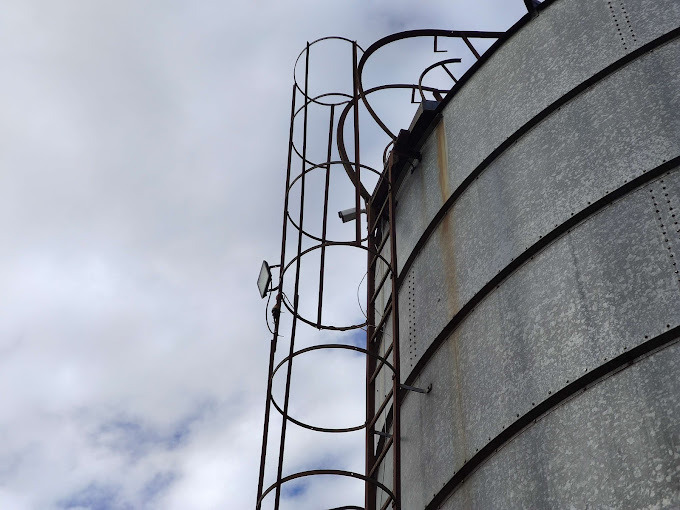 Zewnętrzna kamera monitoringu zamontowana na metalowej konstrukcji z drabinką, na tle pochmurnego nieba, widok z dołu, specyficzny kąt ujęcia.