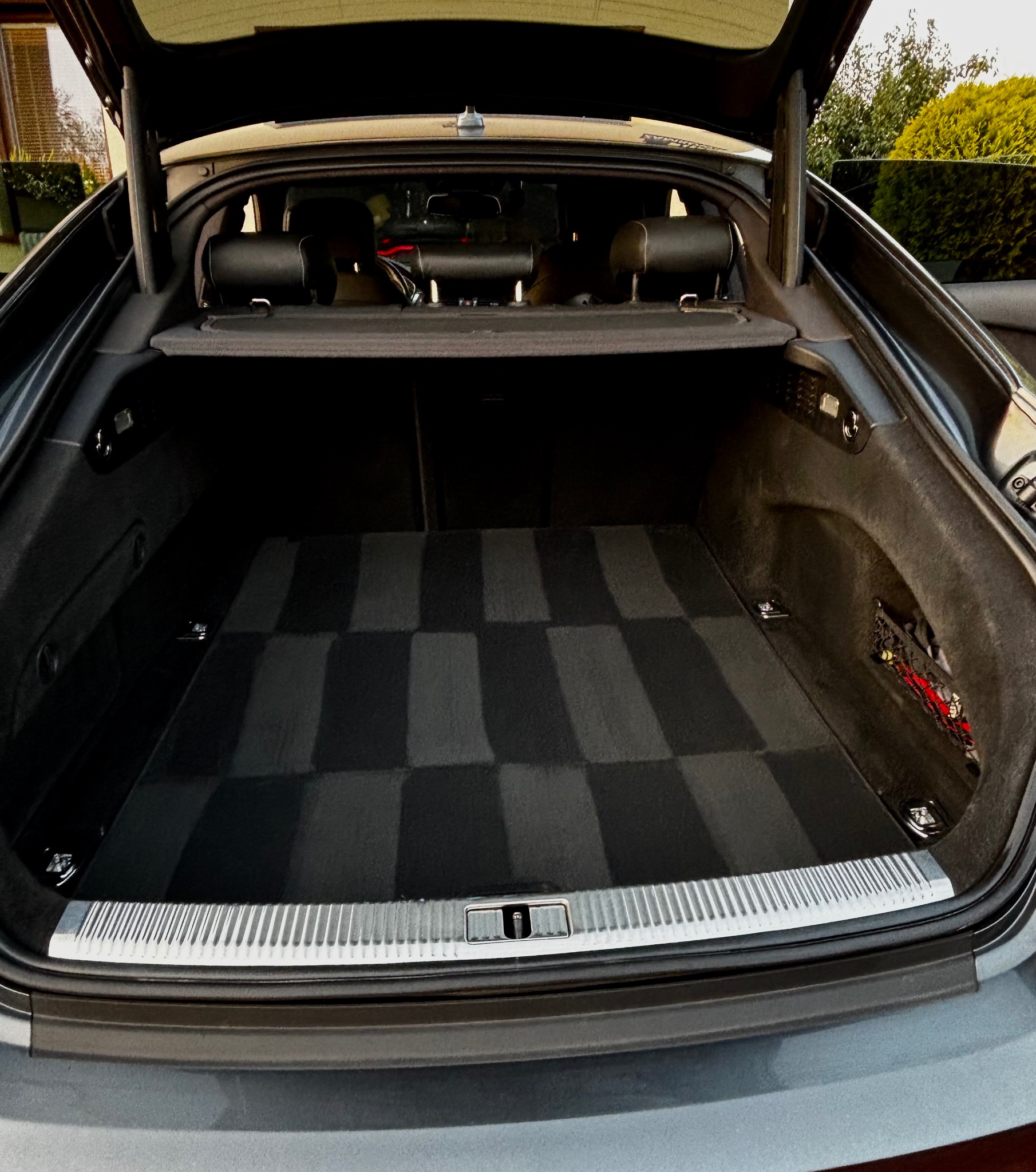 Otwarty bagażnik szarego samochodu z dywanikiem w szachownicę. Widoczne wnętrze bagażnika i fragment karoserii. Na zewnątrz zieleń.