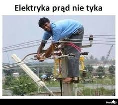 Mężczyzna na słupie energetycznym, naprawiający przewody elektryczne. Widok na otoczenie z zielenią. Ujęcie z dołu, podkreślające wysokość pracy.