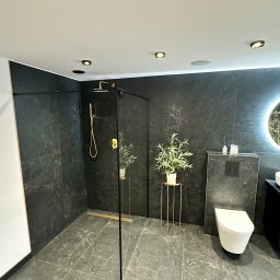 Lucas Dołhań - Elegancka łazienka z kabiną prysznicową typu walk-in, czarnymi płytkami imitującymi marmur, złotą armaturą i rośliną w złotej doniczce. Nowoczesne oświetlenie.