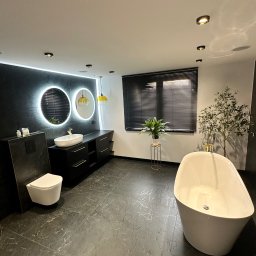 Lucas Dołhań - Nowoczesna łazienka z czarną marmurową podłogą, białą wanną wolnostojącą i podświetlanymi lustrami. Minimalistyczny design z roślinami doniczkowymi.
