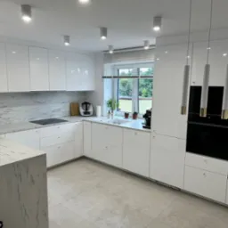 Nowoczesna, jasna kuchnia z białymi, lakierowanymi frontami, marmurowym blatem i wyspą. Minimalistyczne oświetlenie punktowe i wiszące lampy. Okno z widokiem na zieleń.