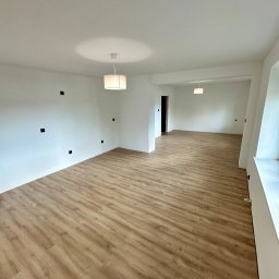 Lucas Dołhań - Jasne, przestronne wnętrze po remoncie: panele podłogowe w odcieniach drewna, białe ściany z widocznymi puszkami elektrycznymi i nowoczesne lampy.