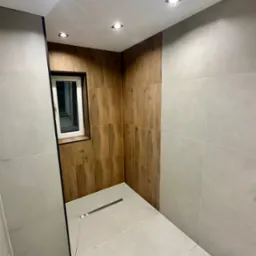 Nowoczesna łazienka po remoncie z drewnopodobnymi i szarymi płytkami, oświetleniem punktowym i małym oknem. Widoczny odpływ liniowy w podłodze.