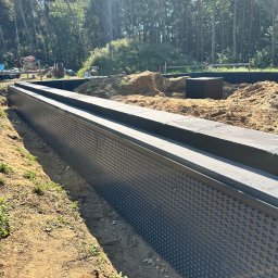 Lucas Dołhań - Ochrona fundamentów budynku z folią kubełkową i betonową nadstawką, widok na wykop, ziemię i las w tle. Prace ziemne i izolacyjne w toku.