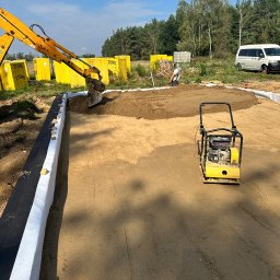 Lucas Dołhań - Żółta koparka rozsypuje ziemię na przygotowany teren z obrzeżami. Obok stoi zagęszczarka, a w tle widoczne palety z bloczkami Ytong i biały bus.