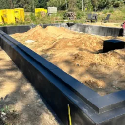 Fundamenty domu jednorodzinnego z izolacją przeciwwilgociową, widoczne bloczki Ytong, ziemia wewnątrz, drzewa w tle. Żółta poziomica oparta o ścianę fundamentu.