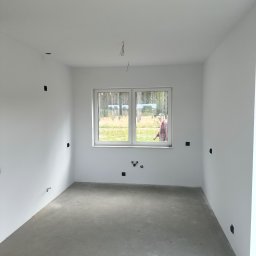 Lucas Dołhań - Surowe wnętrze pokoju z białymi ścianami, szarą podłogą i oknem. Widoczne wystające kable i puszki elektryczne. Osoba w oknie w tle. Stan deweloperski przed wykończeniem.