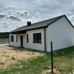 Biały, parterowy dom z czarną dachówką i stolarką okienną na działce. Widoczne fragmenty trawnika i nieba z chmurami. Minimalistyczna architektura.