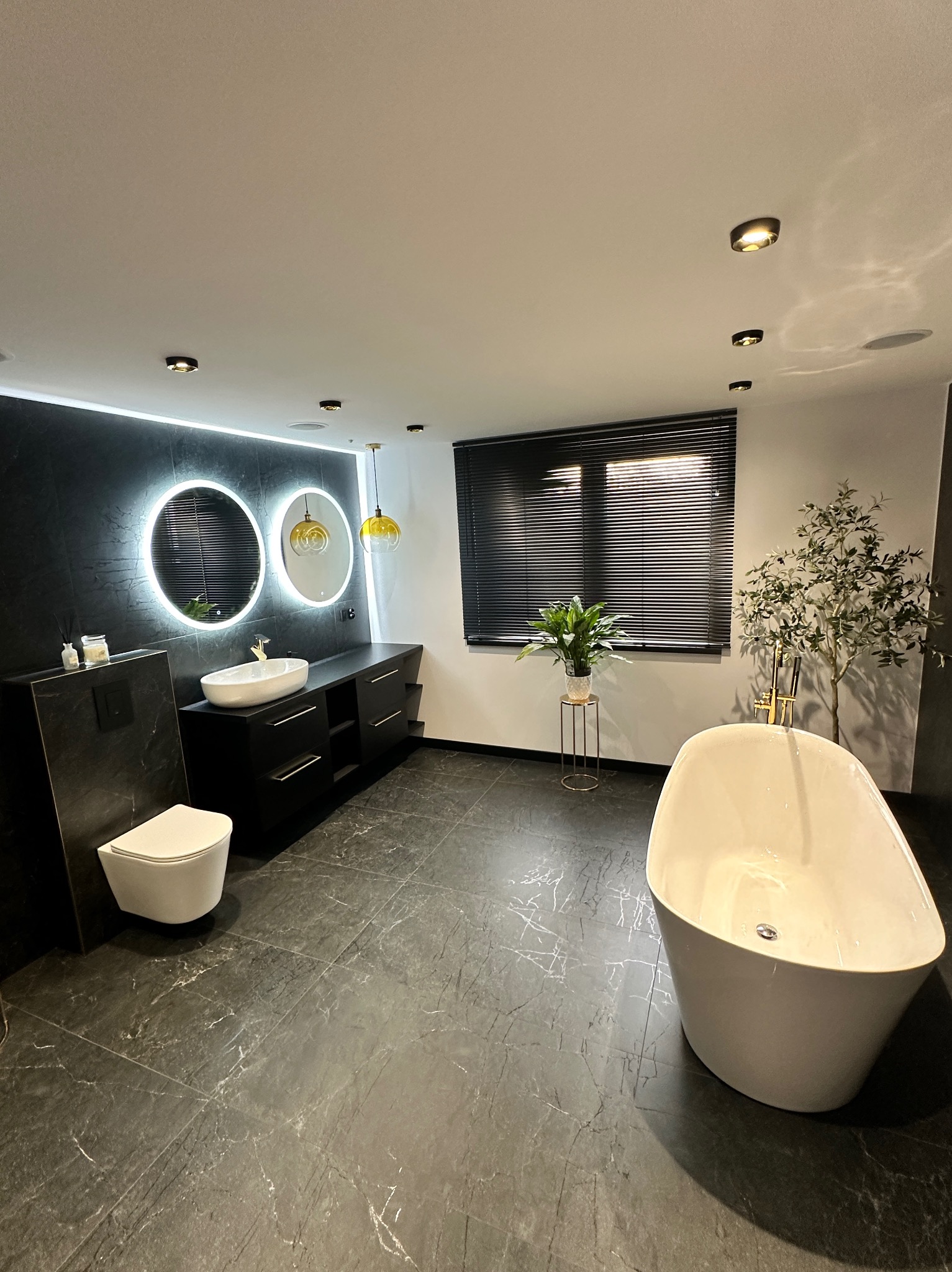 Nowoczesna łazienka z czarną marmurową podłogą, białą wanną wolnostojącą i podświetlanymi lustrami. Minimalistyczny design z roślinami doniczkowymi.