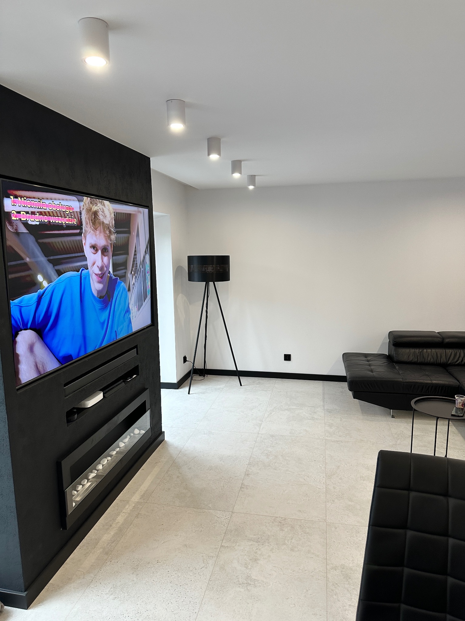 Nowoczesny salon z czarną zabudową TV, bio-kominkiem i minimalistycznym oświetleniem. Beżowe płytki podłogowe i czarne meble tworzą elegancki kontrast.