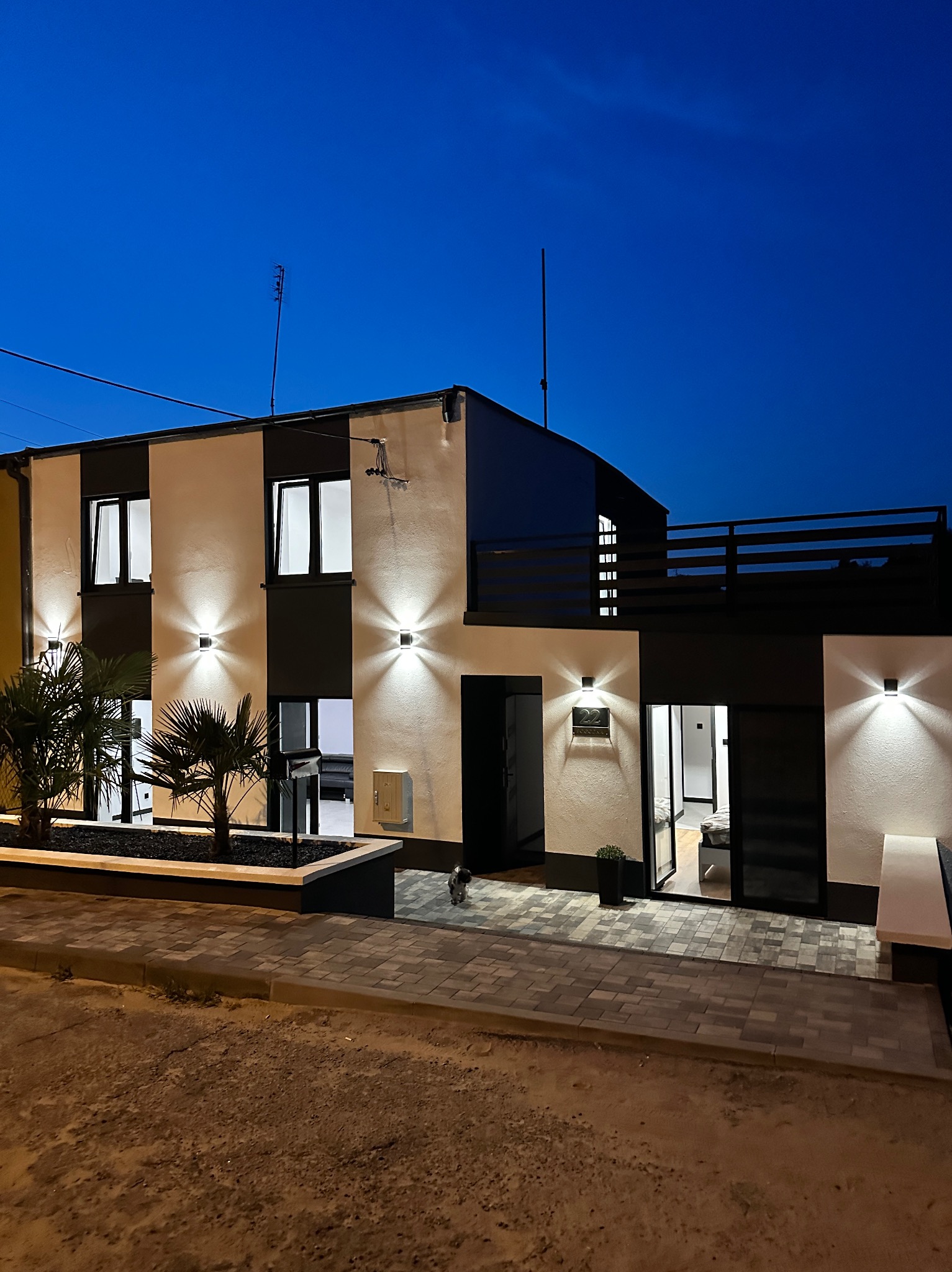 Dwukondygnacyjny budynek po renowacji z czarno-białą elewacją, oświetlony lampami, z roślinami w donicach i kostką brukową przed wejściem, sfotografowany nocą.