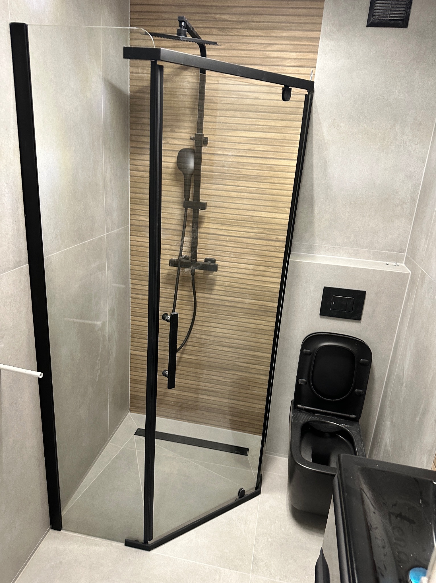 Nowoczesna łazienka po remoncie: kabina prysznicowa z czarnymi ramami, podwieszana toaleta w kolorze czarnym i płytki imitujące drewno na ścianie prysznica.