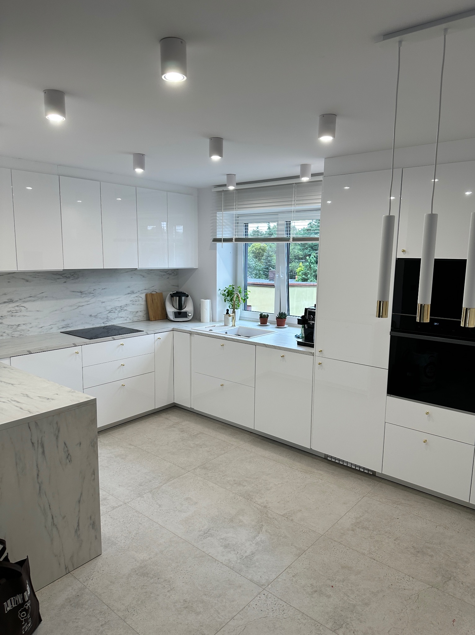 Nowoczesna, jasna kuchnia z białymi, lakierowanymi frontami, marmurowym blatem i wyspą. Minimalistyczne oświetlenie punktowe i wiszące lampy. Okno z widokiem na zieleń.