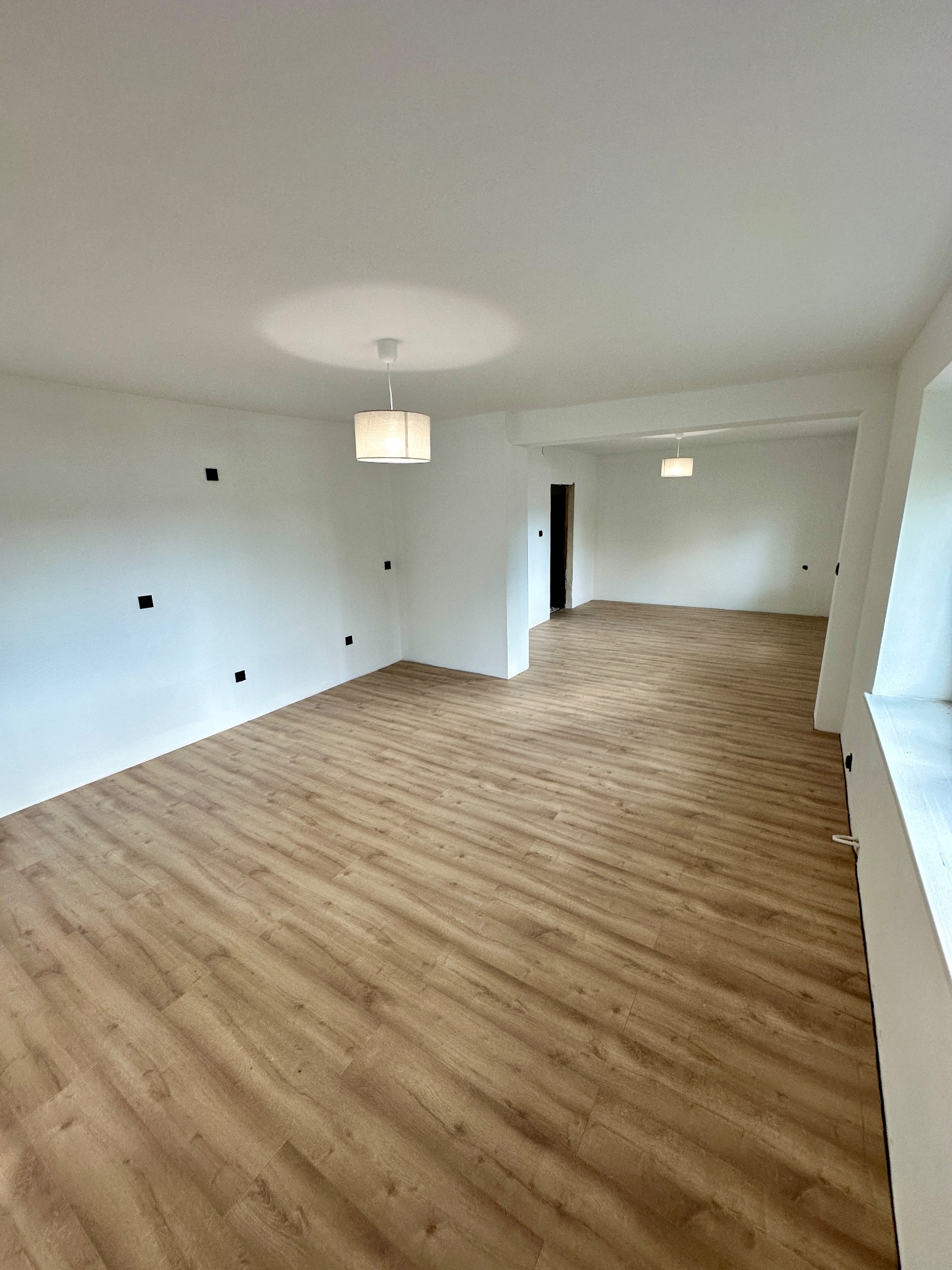 Jasne, przestronne wnętrze po remoncie: panele podłogowe w odcieniach drewna, białe ściany z widocznymi puszkami elektrycznymi i nowoczesne lampy.