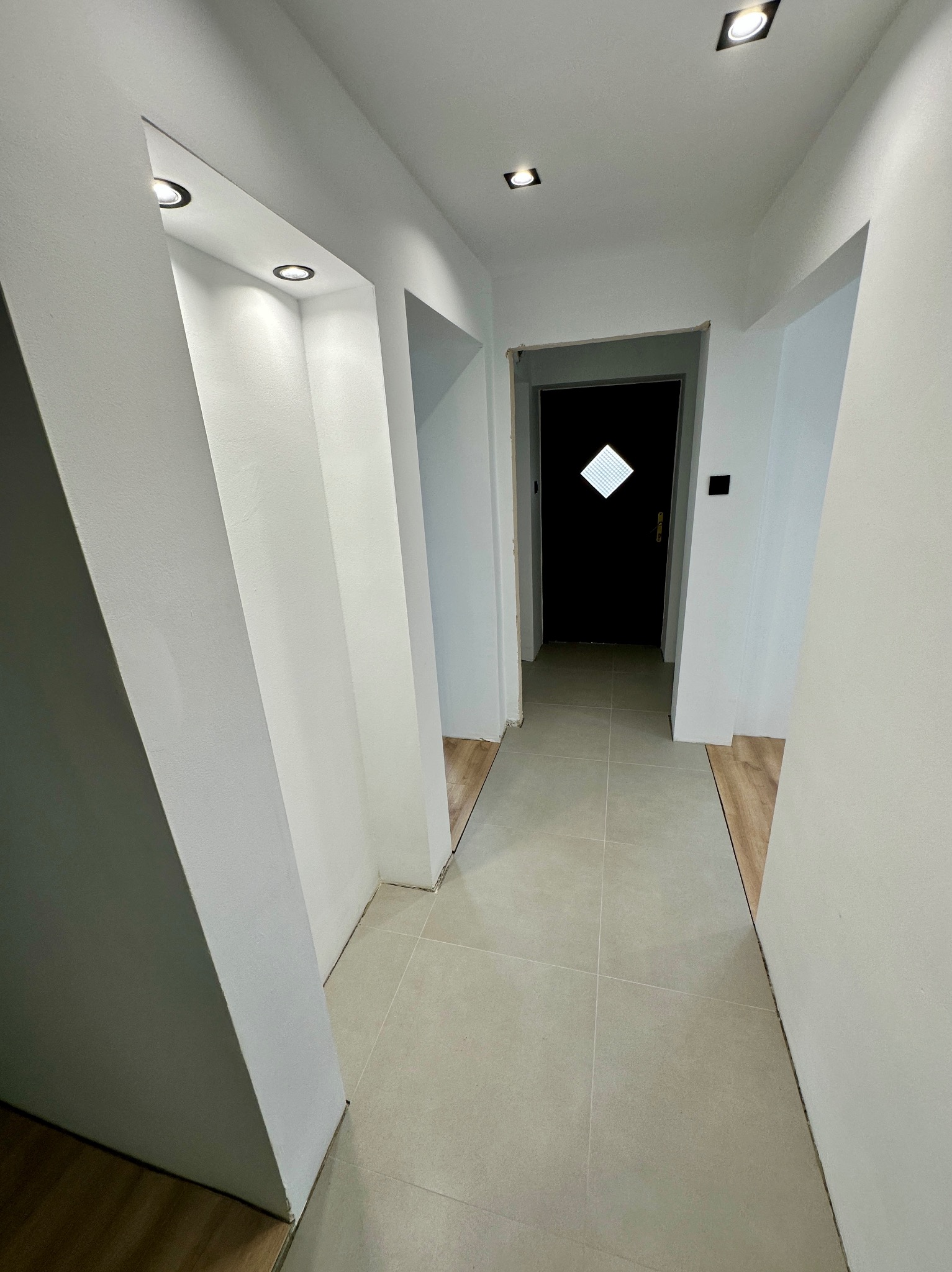 Jasny korytarz po remoncie z białymi ścianami, szarą podłogą z płytek i czarnymi drzwiami. Widoczne nowe oświetlenie sufitowe i fragmenty podłogi przed położeniem płytek.