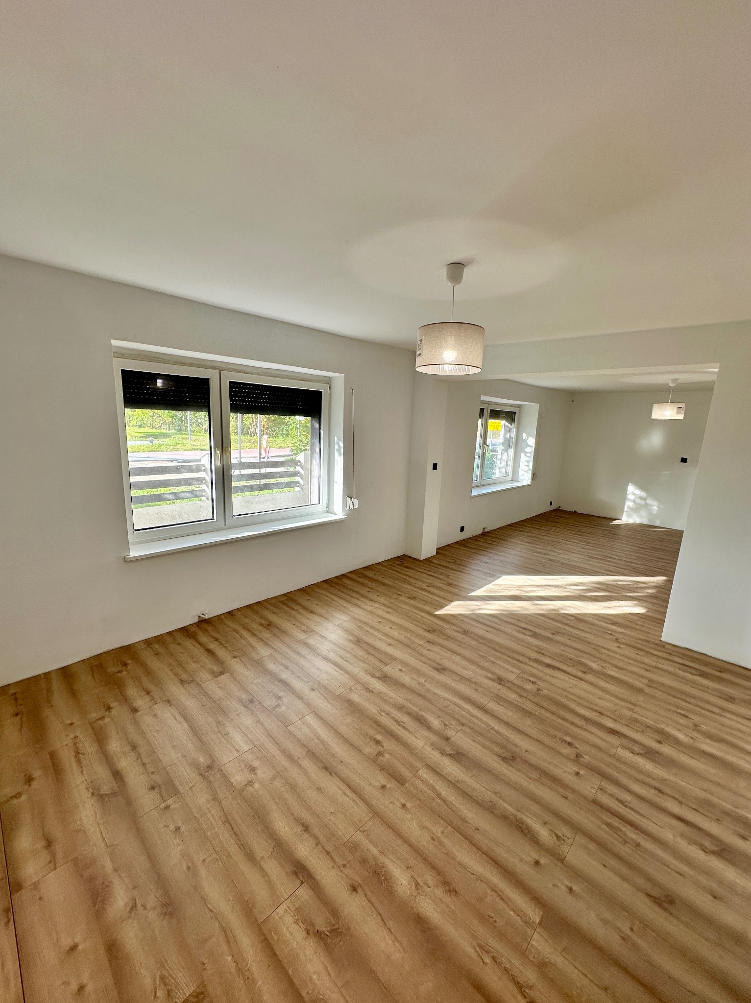 Jasne, przestronne wnętrze po remoncie: panele podłogowe w odcieniach drewna, białe ściany, okna z roletami i nowoczesne lampy sufitowe. Wpadające promienie słoneczne.