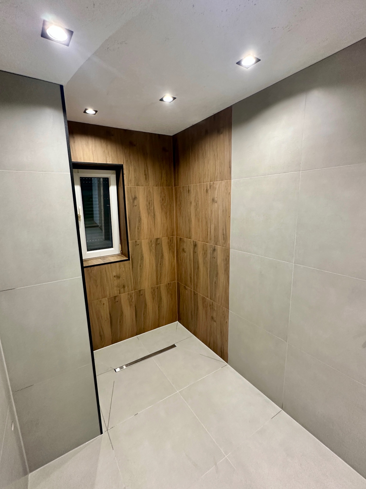 Nowoczesna łazienka po remoncie z drewnopodobnymi i szarymi płytkami, oświetleniem punktowym i małym oknem. Widoczny odpływ liniowy w podłodze.