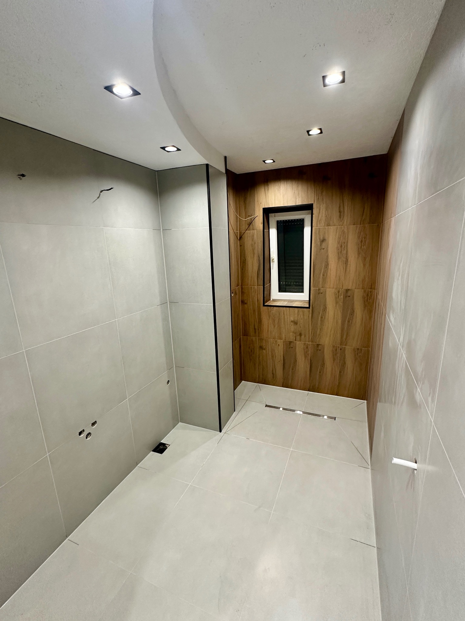 Wykończona łazienka z szarymi i drewnopodobnymi płytkami. Widoczne odpływ liniowy, okno z czarną ramą i oświetlenie sufitowe.