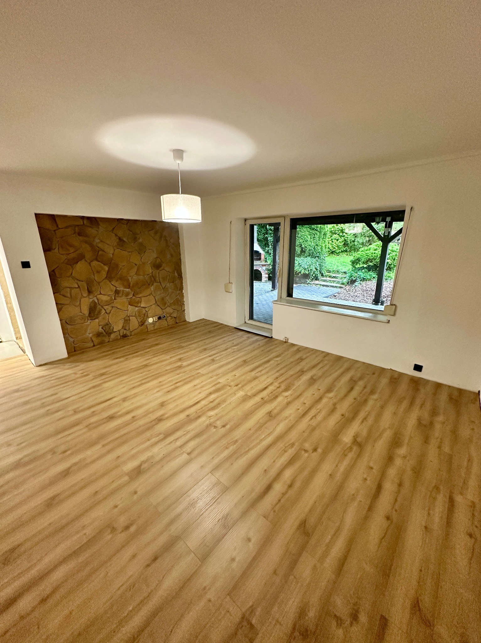 Jasna podłoga drewnopodobna w pustym pokoju z oknem na ogród i ścianą z kamienia dekoracyjnego. Nowoczesna lampa sufitowa. Widok na zewnątrz przez otwarte drzwi.