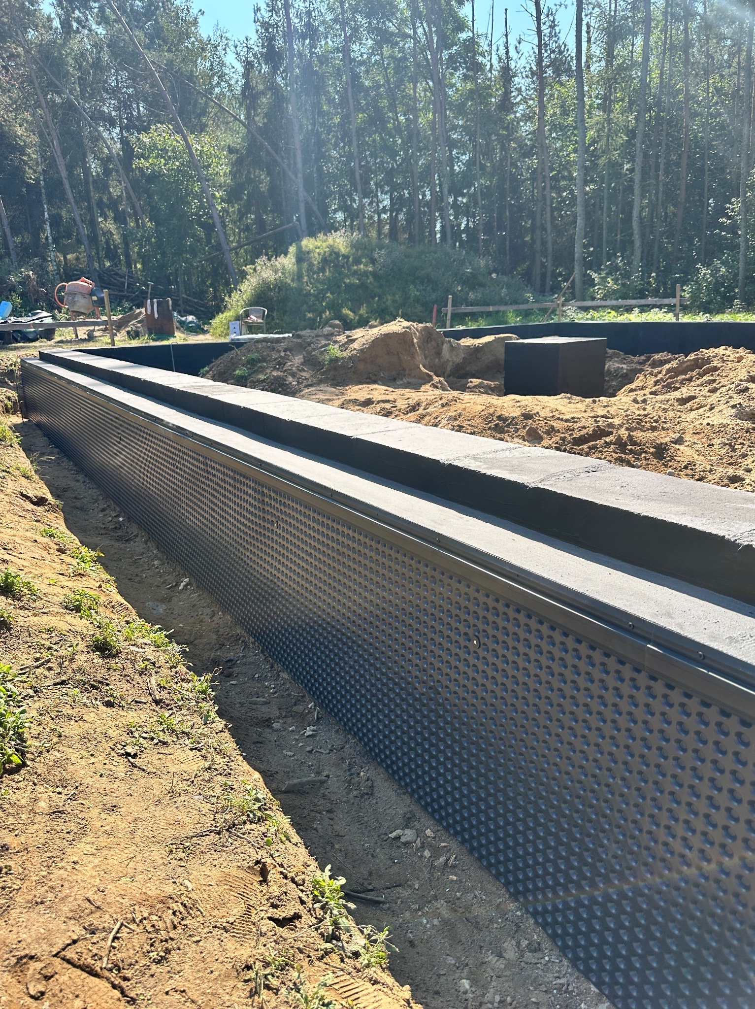 Ochrona fundamentów budynku z folią kubełkową i betonową nadstawką, widok na wykop, ziemię i las w tle. Prace ziemne i izolacyjne w toku.