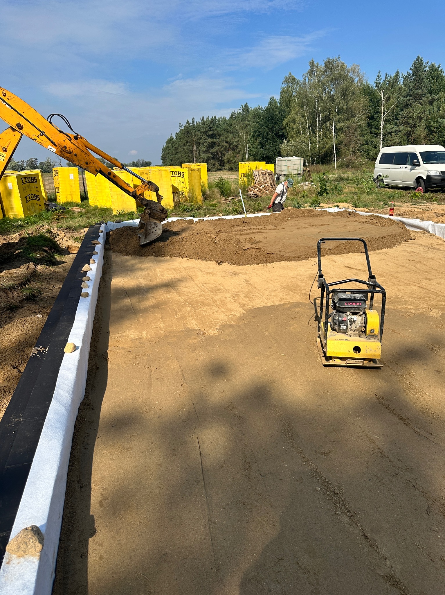 Żółta koparka rozsypuje ziemię na przygotowany teren z obrzeżami. Obok stoi zagęszczarka, a w tle widoczne palety z bloczkami Ytong i biały bus.