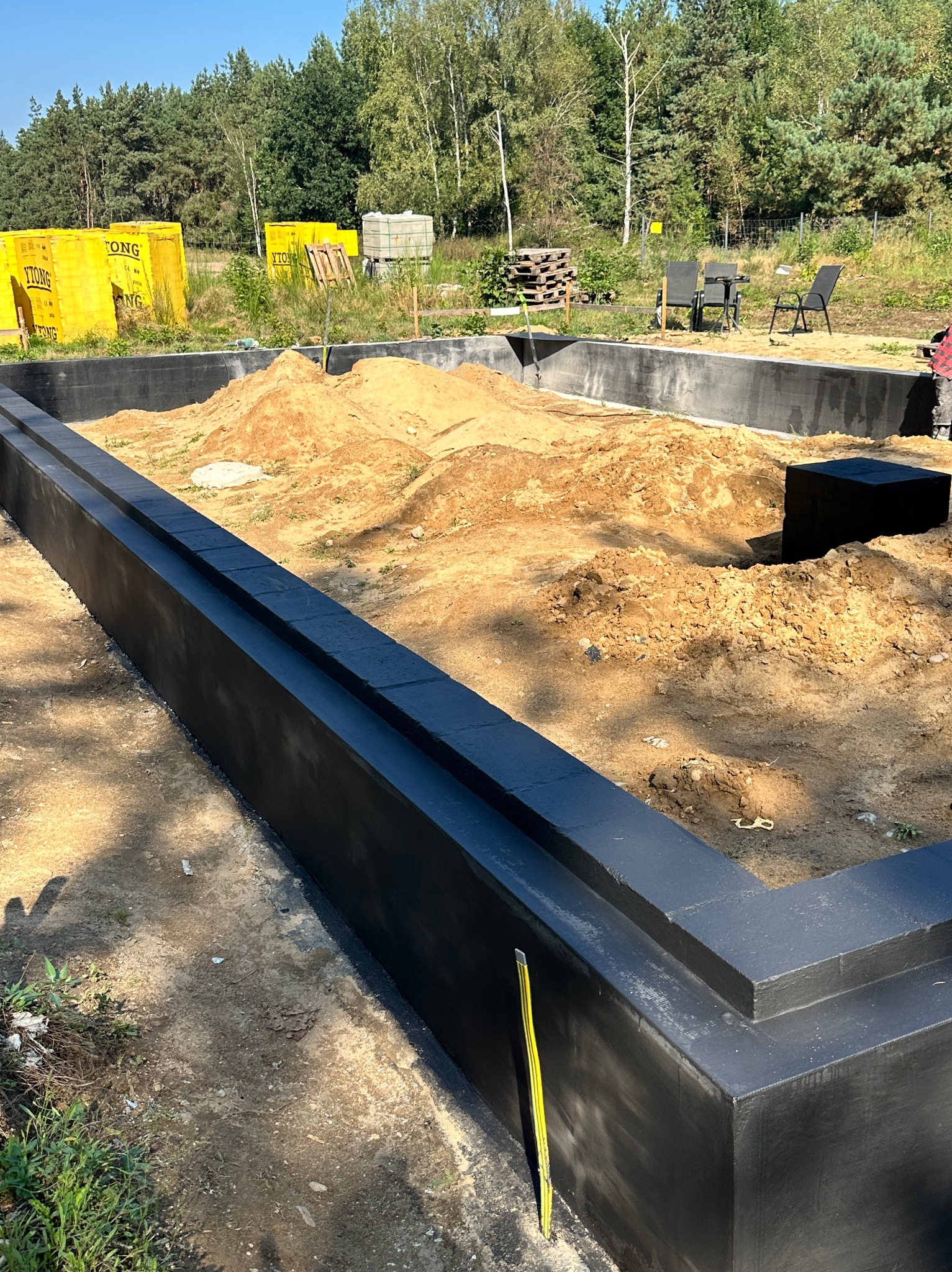 Fundamenty domu jednorodzinnego z izolacją przeciwwilgociową, widoczne bloczki Ytong, ziemia wewnątrz, drzewa w tle. Żółta poziomica oparta o ścianę fundamentu.